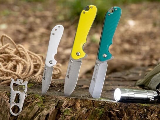 3 folding pocket knives with blades stuck slightly into the ground and shown with a steel multi tool and a mini led flashlight